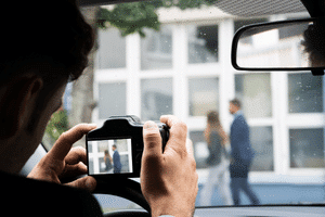 A private investigator in a car using a camera to surveil a couple walking in front of a building as the subjects of a sub rosa investigation.
